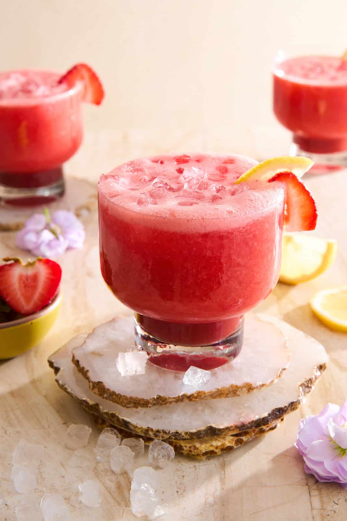 A glass of pink strawberry lemonade with ice, garnished with a lemon slice and strawberry, sits on stone coasters.