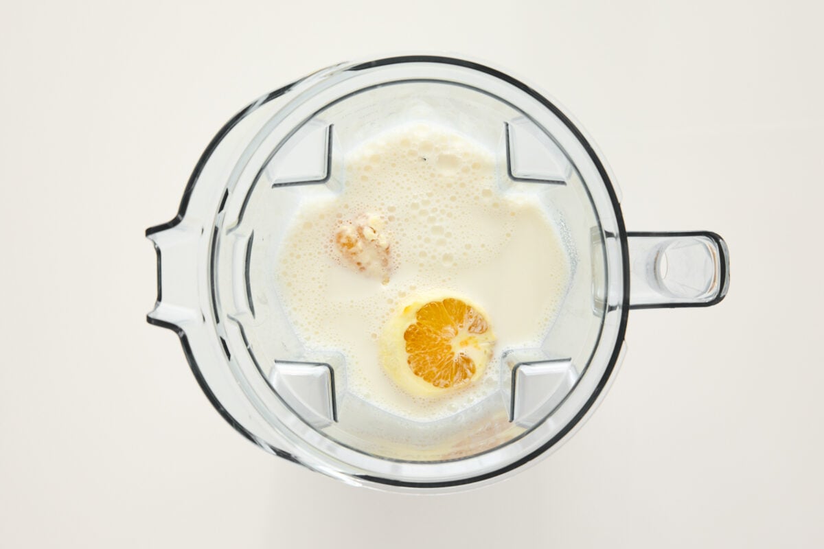 Top-down view of a blender containing milk, an orange half, and other ingredients, ready to be blended, on a white background.