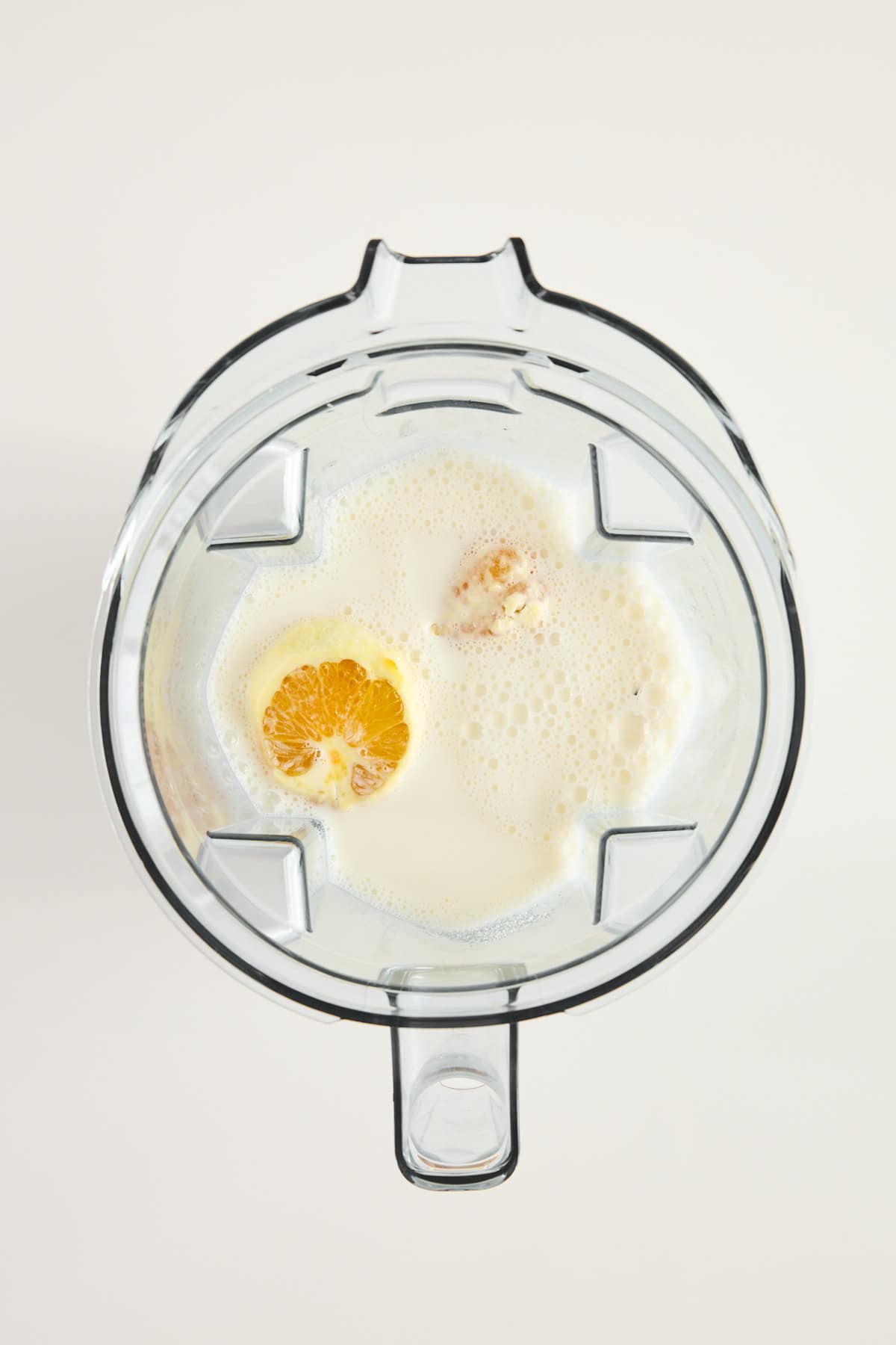 Top-down view of a blender containing milk, an orange half, and other ingredients, ready to be blended, on a white background.