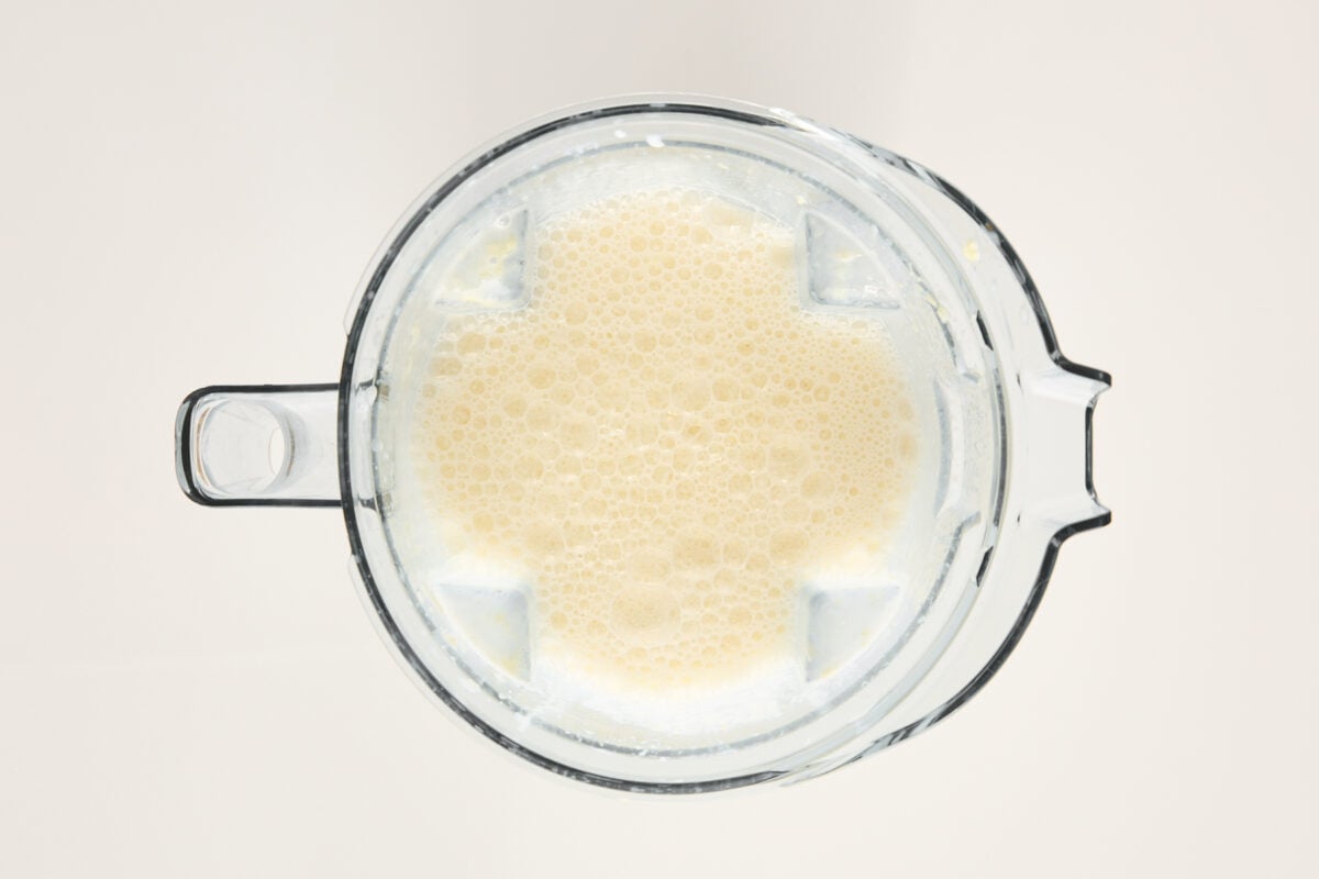 A top-down view of a blender filled with a light beige, frothy liquid, likely a smoothie or blended beverage, against a plain light background.