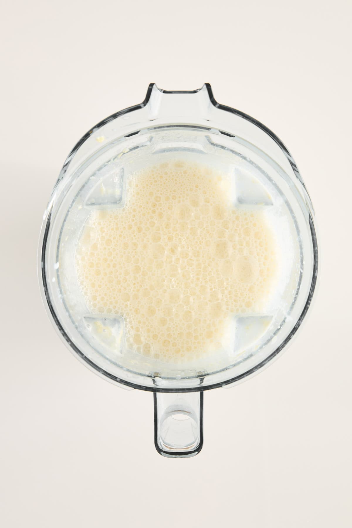 A top-down view of a blender filled with a light beige, frothy liquid, likely a smoothie or blended beverage, against a plain light background.
