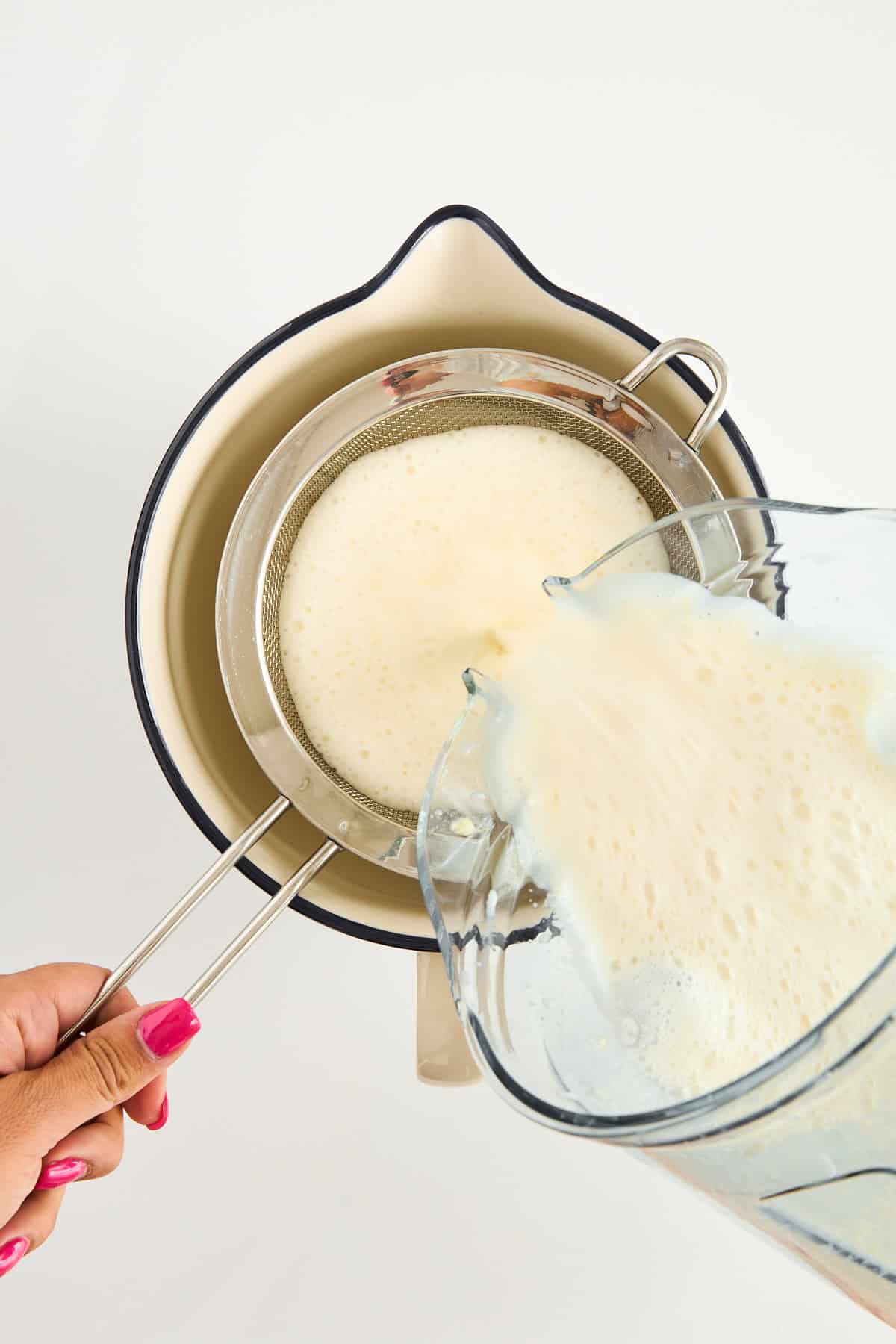A hand holds a metal strainer over a cream-colored bowl while pouring a frothy liquid from a blender through the strainer. The person has pink nail polish. The background is white.