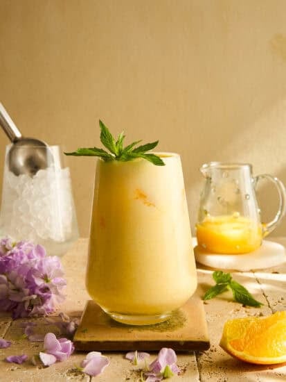 A creamy orange smoothie garnished with a sprig of mint sits on a tile coaster, with a small pitcher of orange juice, an ice scoop, purple flowers, and an orange wedge nearby.