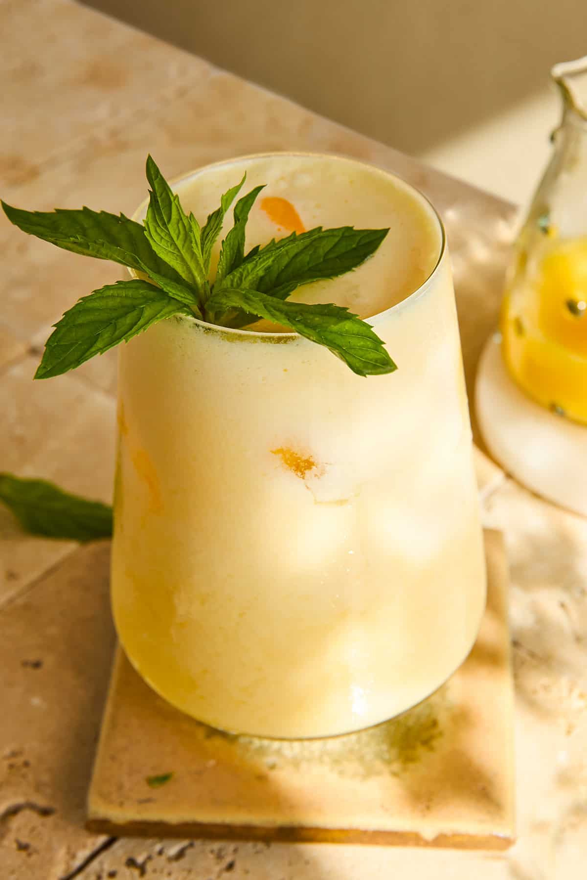 A frosty yellow drink garnished with a sprig of fresh mint sits on a square tile coaster, with sunlight casting soft shadows on the stone surface. A glass pitcher with yellow liquid is partly visible in the background.