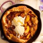 A peach galette with a golden crust sits in a black skillet, topped with two scoops of vanilla ice cream and a drizzle of honey. Slices of fresh peach and a floral napkin are nearby on a light-colored surface.