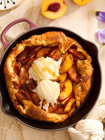 A peach galette with a golden crust sits in a black skillet, topped with two scoops of vanilla ice cream and a drizzle of honey. Slices of fresh peach and a floral napkin are nearby on a light-colored surface.