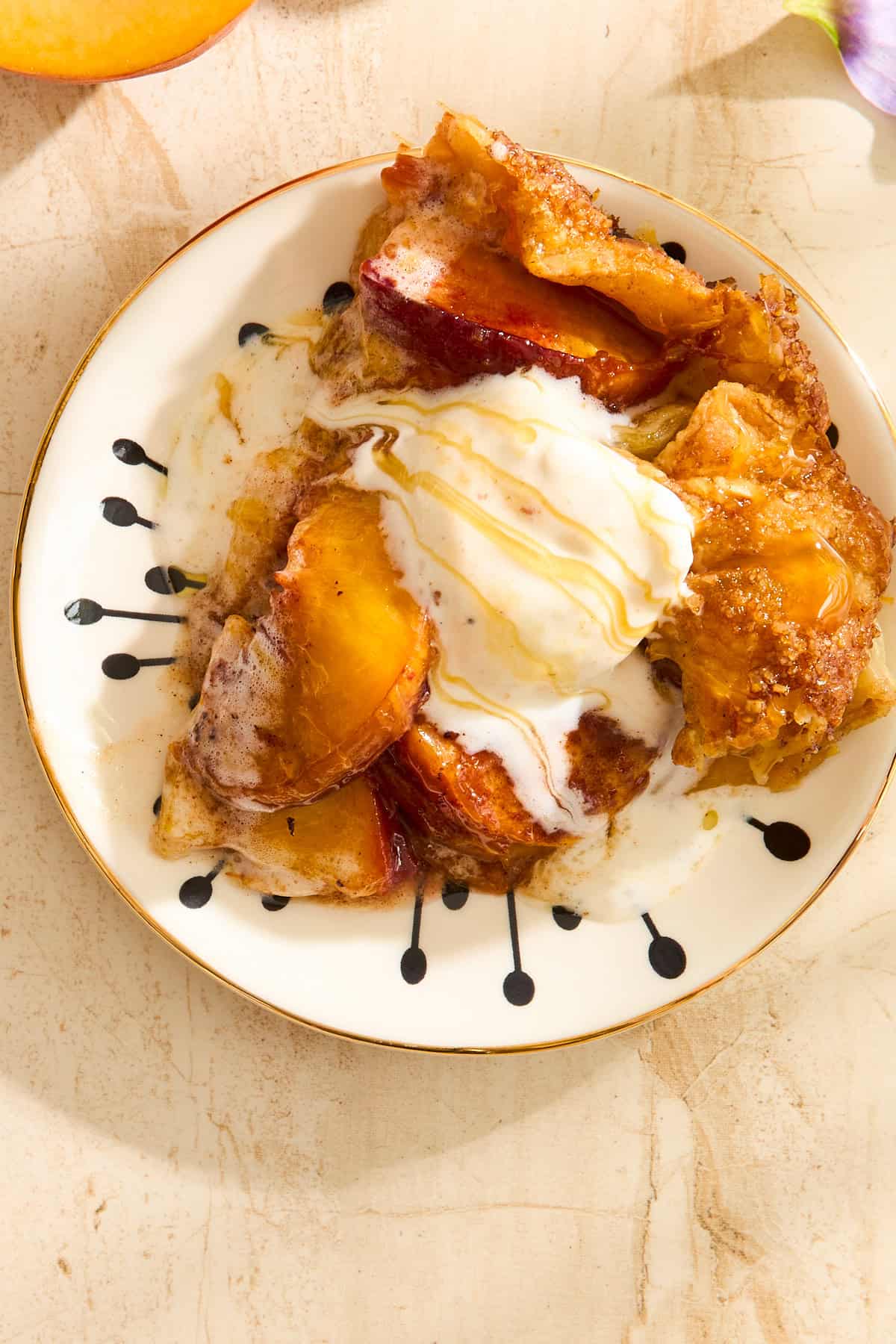 A plate with a serving of peach cobbler topped with melting vanilla ice cream and a drizzle of syrup, on a light-colored surface.