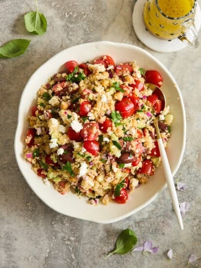 A white bowl filled with a colorful salad of couscous, cherry tomatoes, feta cheese, red onion, and fresh greens, with a white spoon. A small jar of yellow dressing sits nearby on a marble surface.