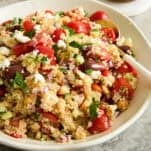 A bowl of quinoa salad with chopped tomatoes, cucumbers, red onions, kalamata olives, parsley, and crumbled feta cheese, with a spoon and a small jar of dressing in the background.