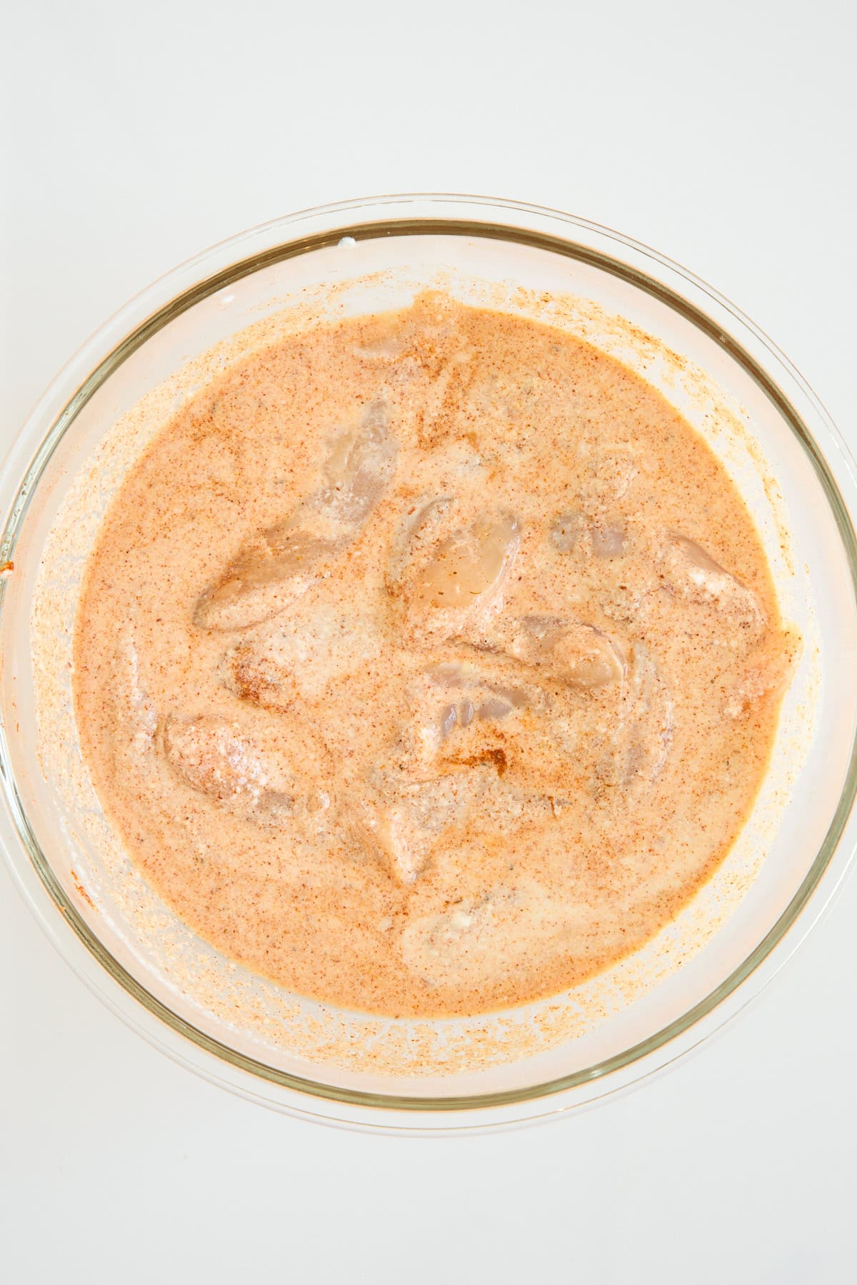 A clear glass bowl filled with pieces of chicken marinating in a creamy, orange-colored spice mixture, set against a plain white background.