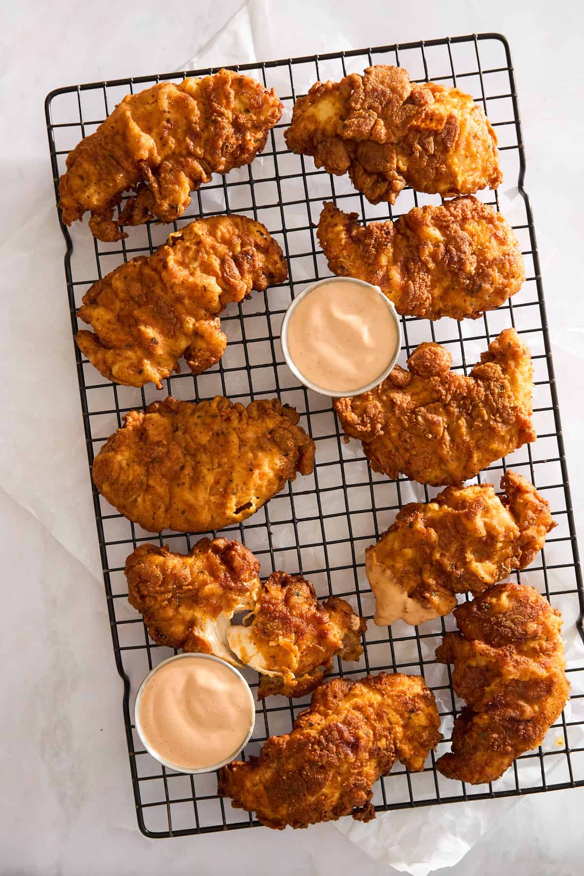 A cooling rack holds several pieces of crispy fried chicken tenders, accompanied by three small cups of creamy dipping sauce. The setup is on a light surface, giving a fresh, appetizing appearance.