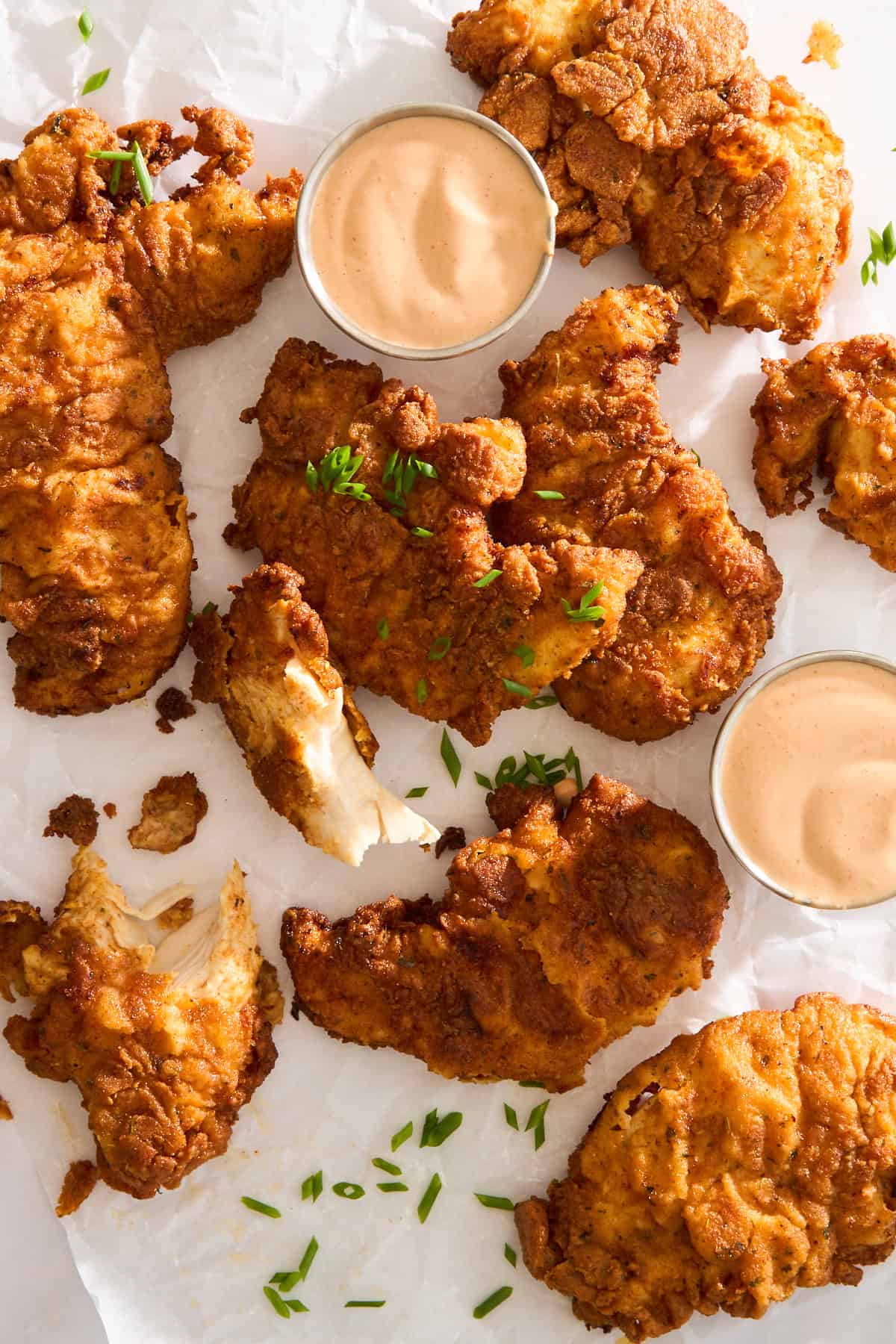 Crispy fried chicken tenders arranged on parchment paper with two ramekins of dipping sauce and a sprinkle of chopped chives.
