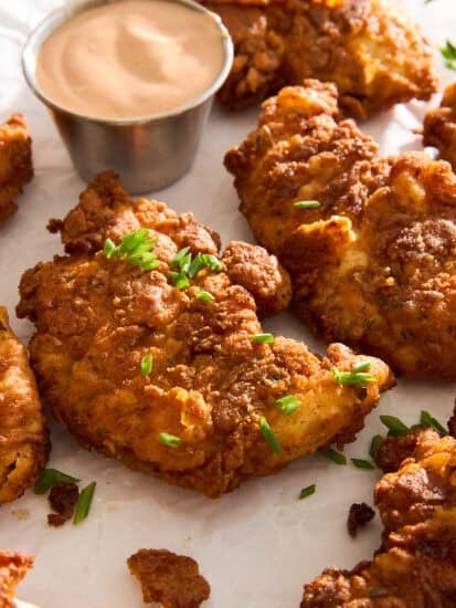 Crispy fried chicken pieces garnished with chopped chives are arranged on parchment paper next to a small metal cup of creamy dipping sauce.
