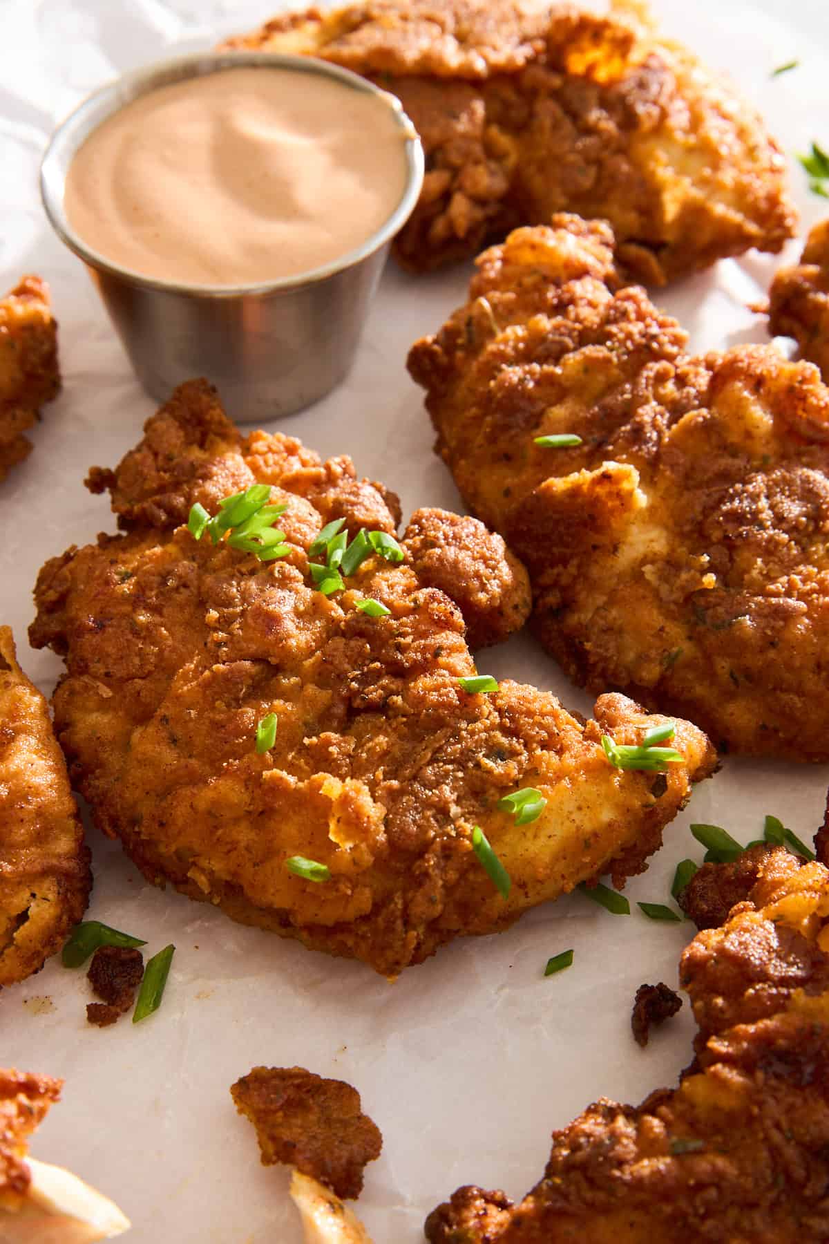 Crispy fried chicken pieces garnished with chopped chives are arranged on parchment paper next to a small metal cup of creamy dipping sauce.
