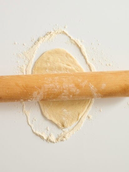 A wooden rolling pin rolls out a circle of dough on a white surface dusted with flour.
