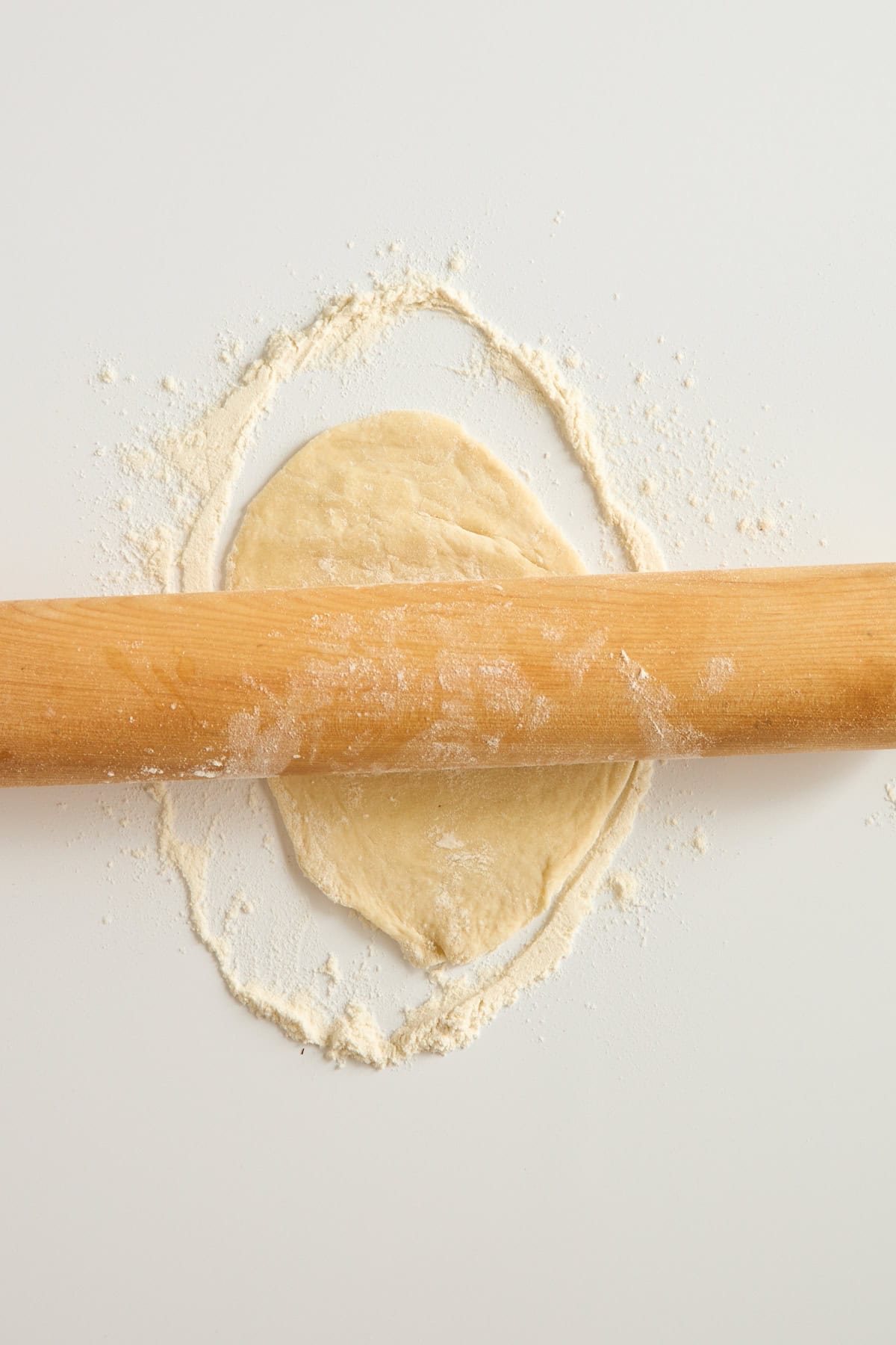 A wooden rolling pin rolls out a circle of dough on a white surface dusted with flour.
