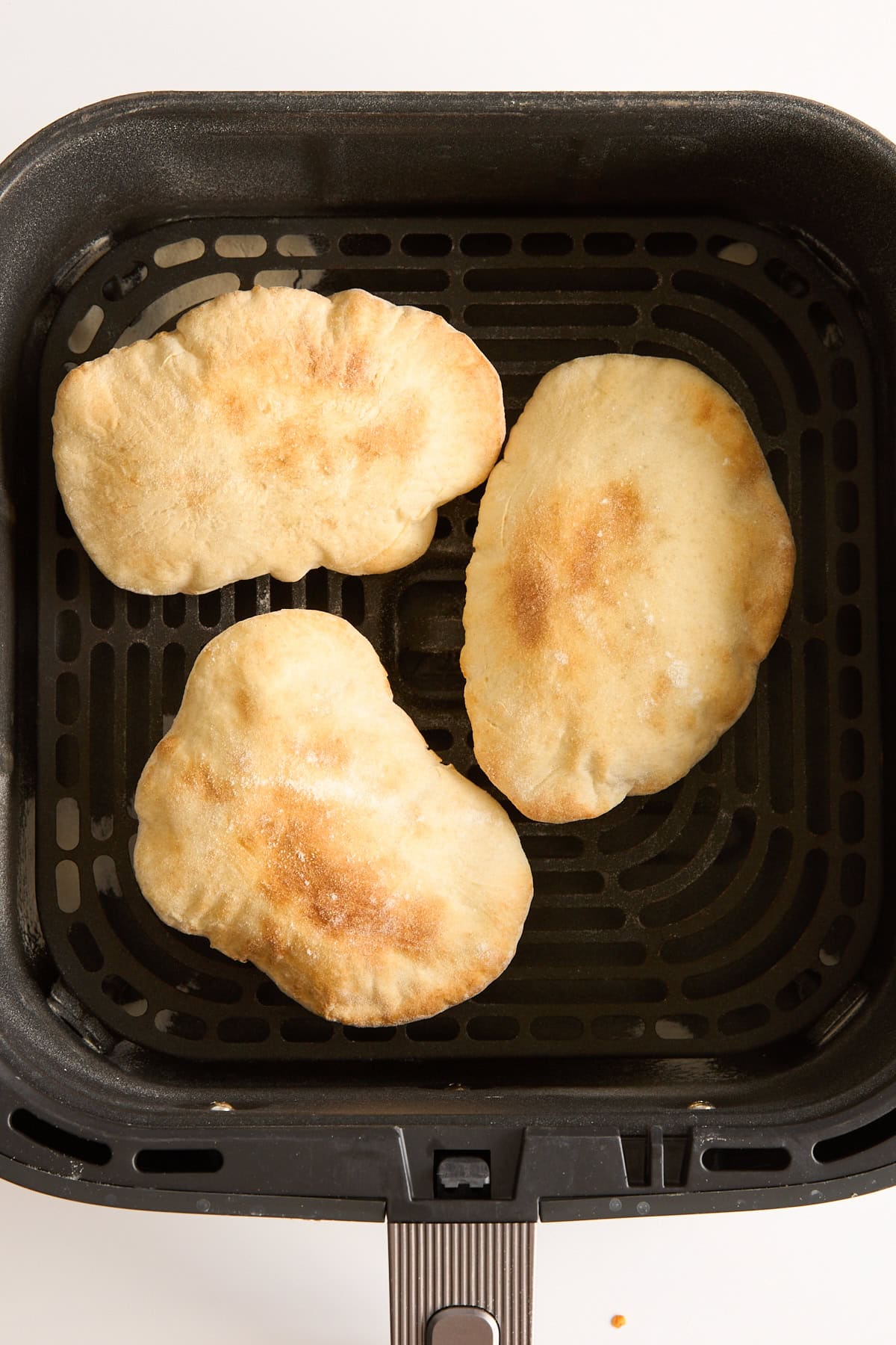 Three pieces of golden-brown flatbread are placed inside the basket of an air fryer, viewed from above on a white background.