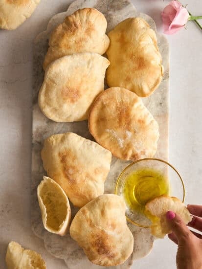 A marble tray topped with several pieces of pita bread sits on a light surface. A hand dips a piece of pita into a small glass bowl of olive oil. Pink roses are visible in the top right corner.