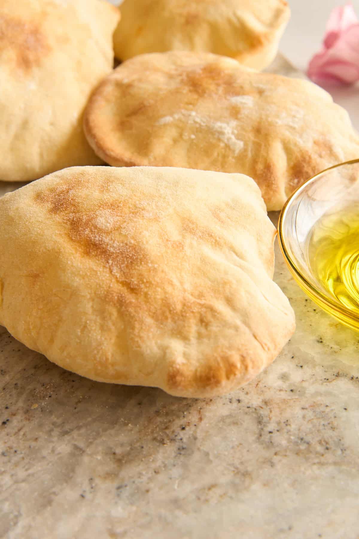 Three pieces of pita bread rest on a light-colored stone surface next to a small glass bowl filled with olive oil.