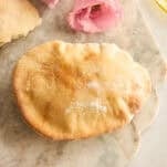 A golden-brown pita bread sits on a marble surface next to two pink flowers and a partial view of a glass dish with olive oil.