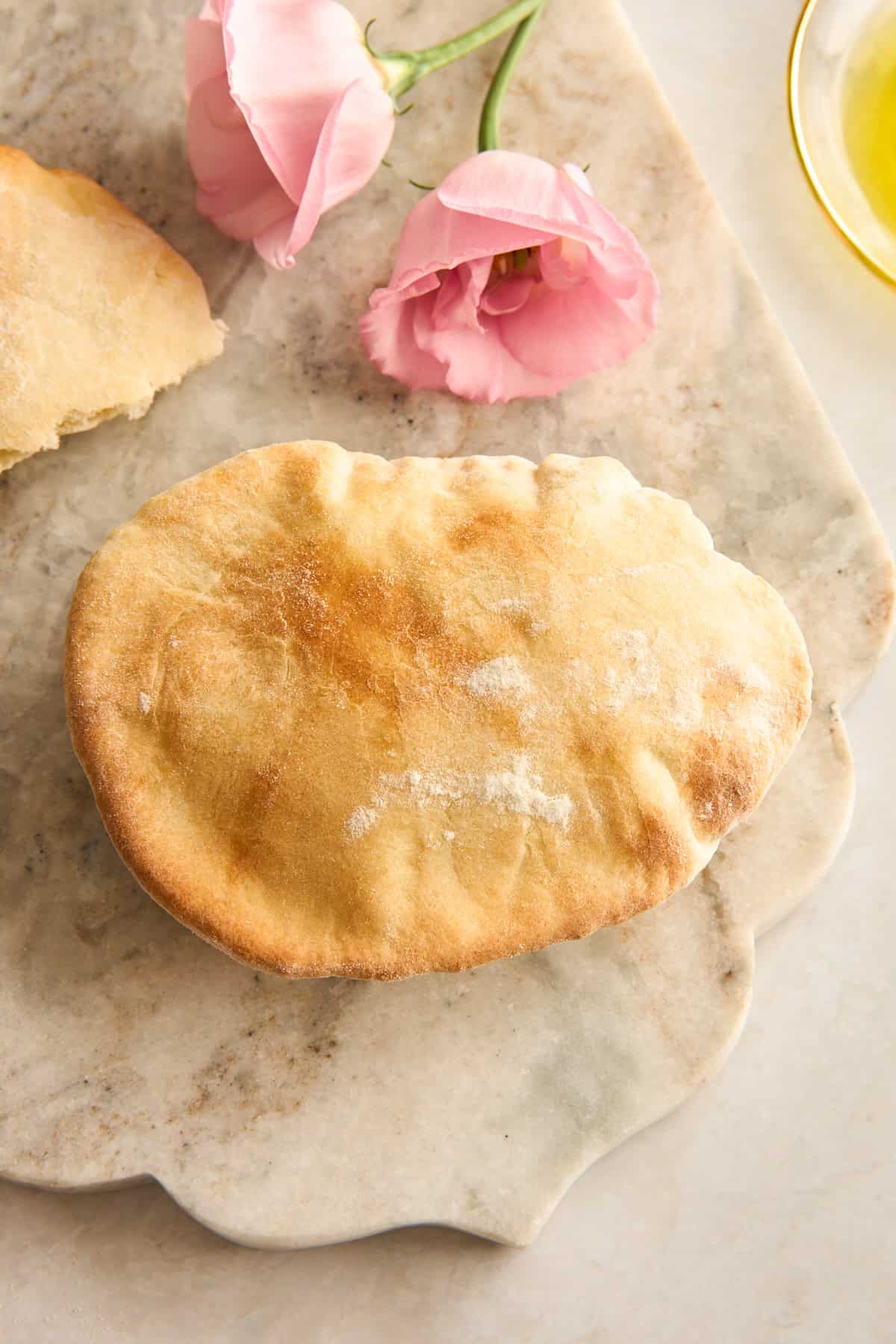 A golden-brown pita bread sits on a marble surface next to two pink flowers and a partial view of a glass dish with olive oil.