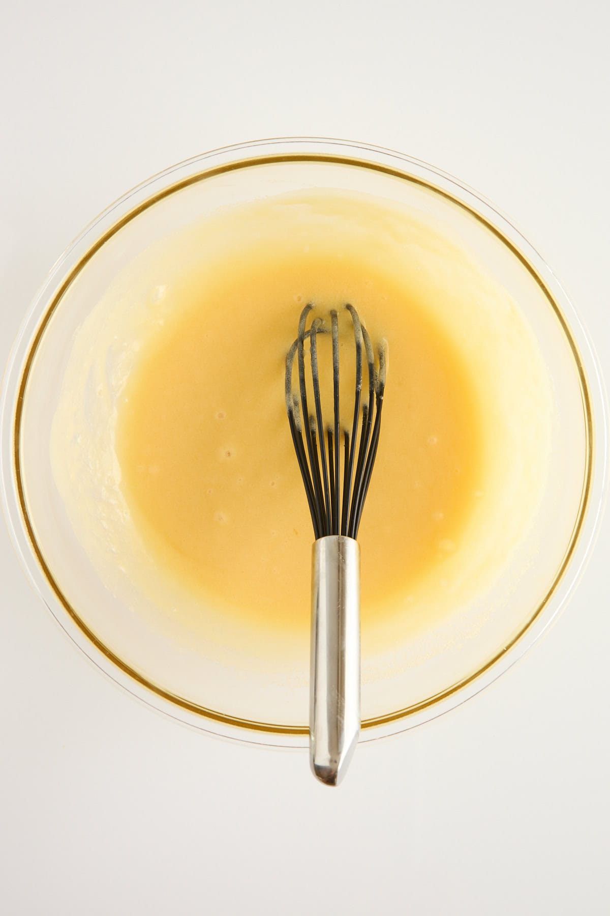A metal whisk in a glass bowl containing pale yellow batter or mixture, set against a white background.