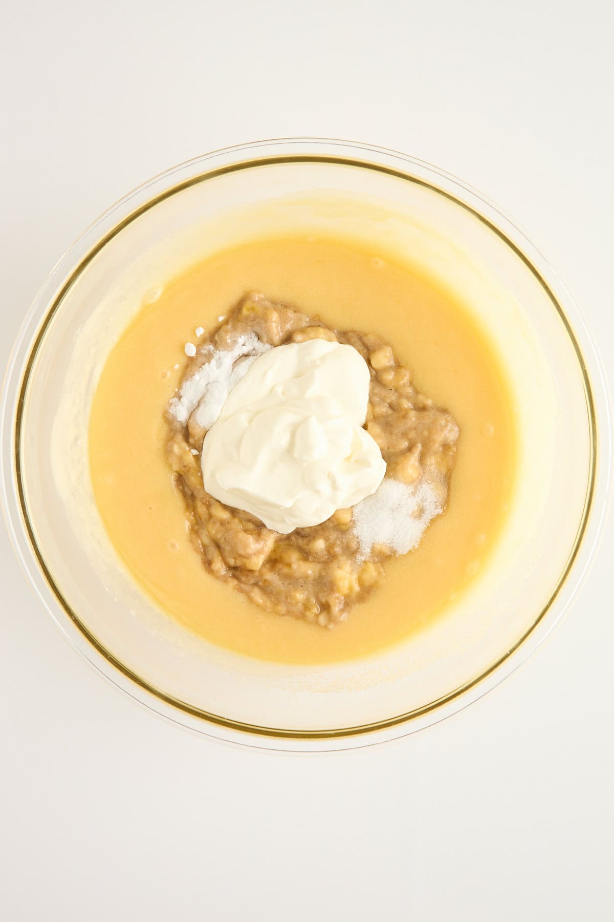 A glass bowl filled with a yellow batter, mashed bananas, white yogurt, and a sprinkle of baking soda, viewed from above on a white surface.