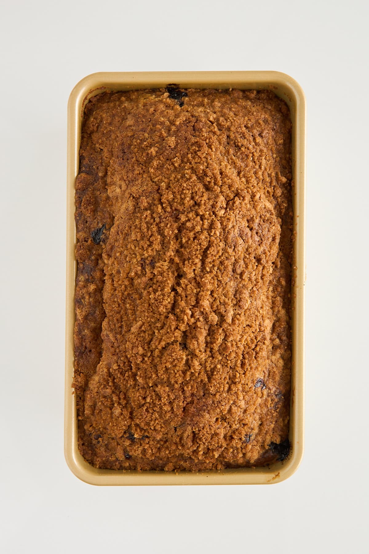 A rectangular loaf of baked bread with a crumbly brown crust is shown in a tan loaf pan, viewed from above on a white background.