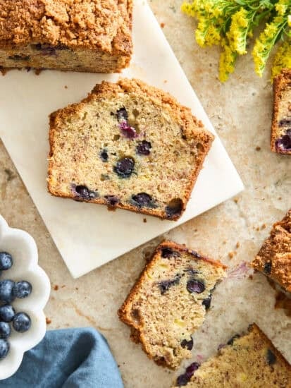 Sliced blueberry bread with a crumb topping is arranged on a marble board. A bowl of fresh blueberries, a blue napkin, and yellow flowers are nearby on a light countertop.