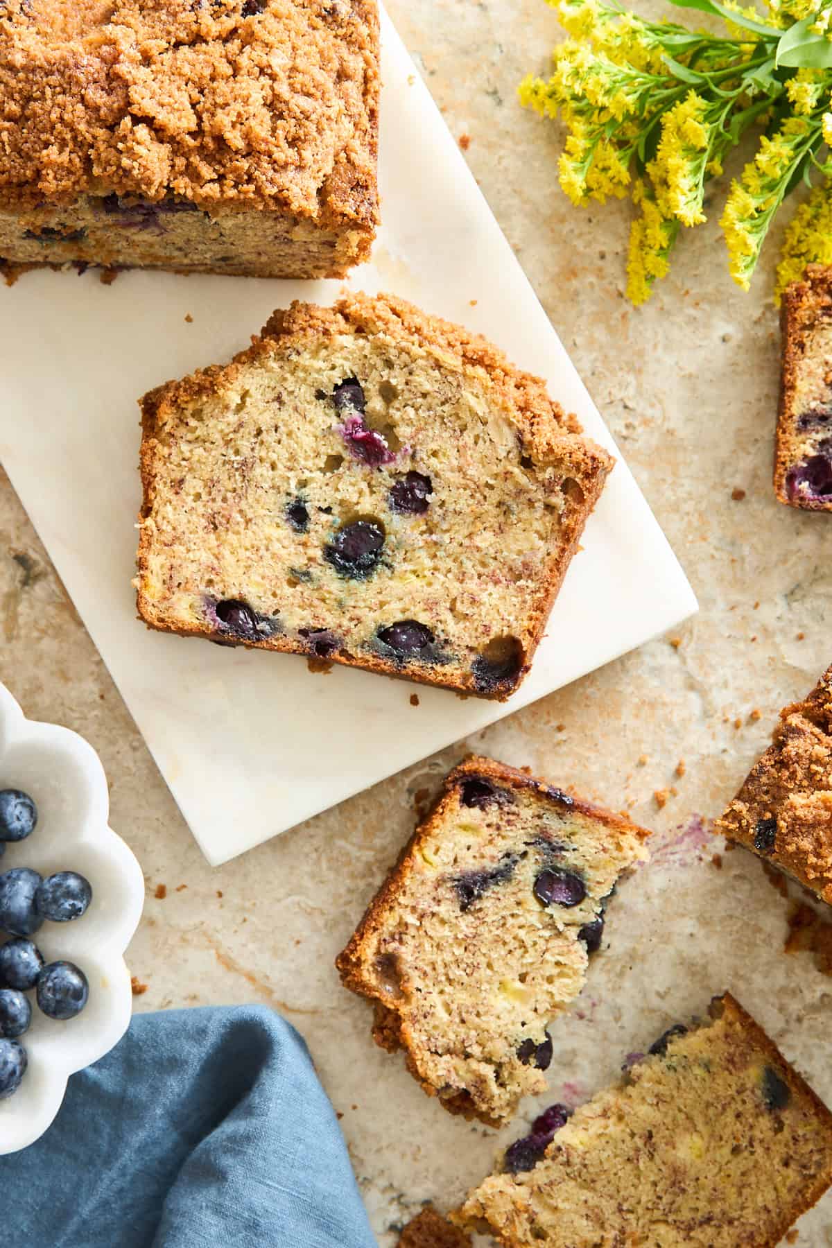 Sliced blueberry bread with a crumb topping is arranged on a marble board. A bowl of fresh blueberries, a blue napkin, and yellow flowers are nearby on a light countertop.
