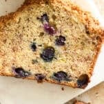 A close-up of a slice of blueberry zucchini bread on a white surface, showing a golden-brown crust and moist interior with visible blueberries and shredded zucchini.