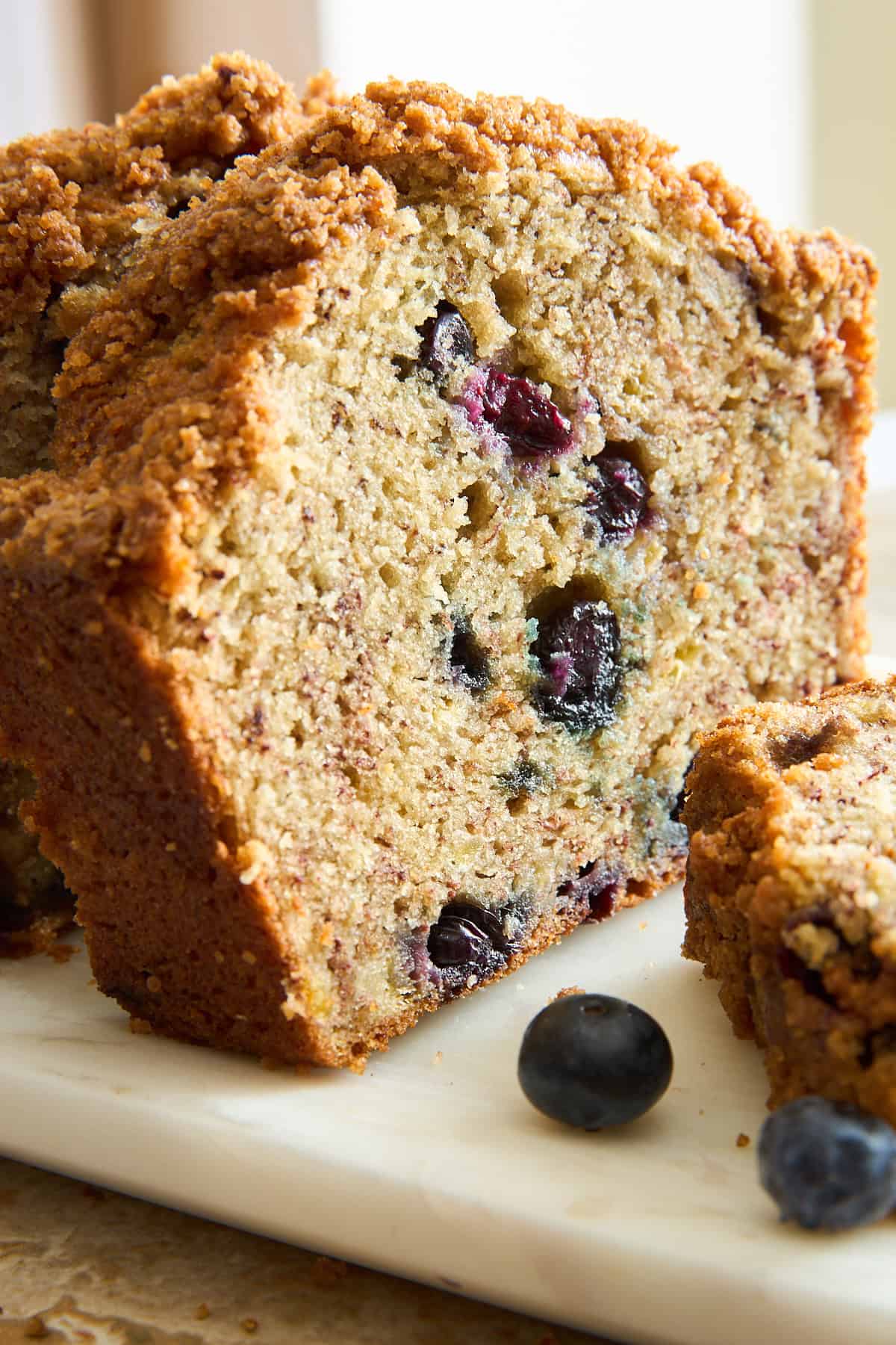 A close-up of a sliced blueberry bread loaf with a crumbly topping, sitting on a white marble surface. Fresh blueberries are visible inside the bread and scattered nearby.