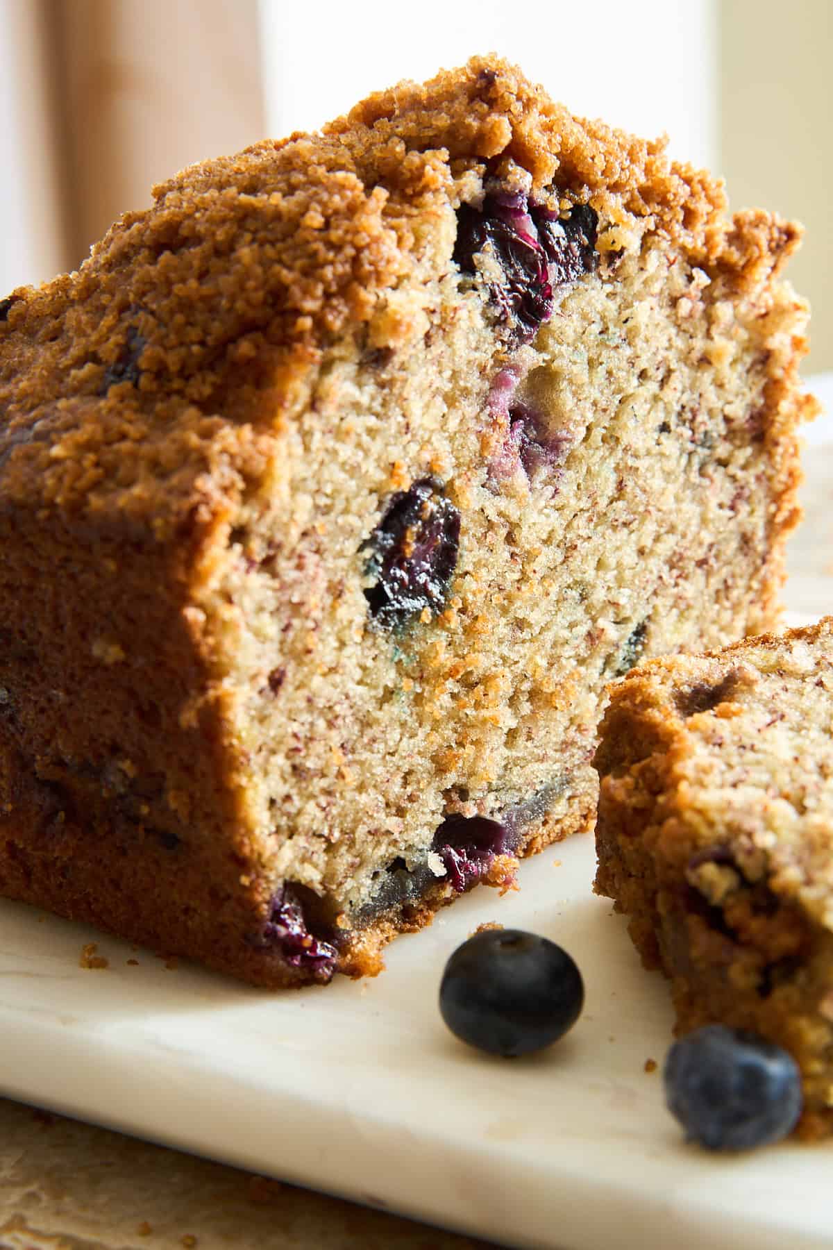 A close-up of a sliced blueberry bread loaf with a crumbly topping, showing moist texture and visible blueberries inside. Two fresh blueberries are placed on the marble surface nearby.