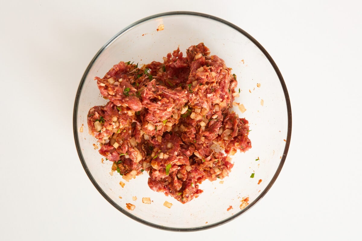 A clear glass bowl filled with a mixture of raw ground meat, chopped onions, herbs, and spices on a white background.
