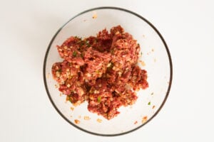 A clear glass bowl filled with a mixture of raw ground meat, chopped onions, herbs, and spices on a white background.