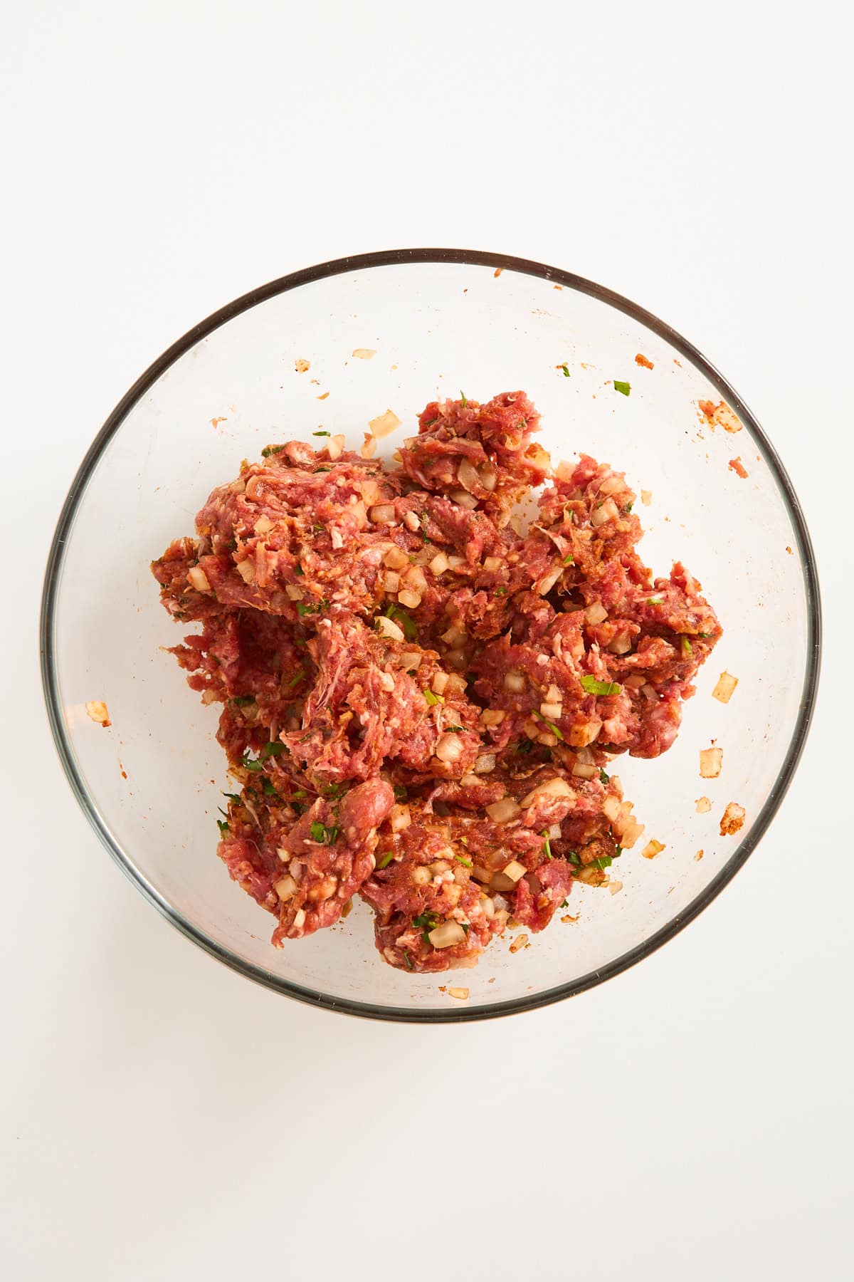 A clear glass bowl filled with a mixture of raw ground meat, chopped onions, herbs, and spices on a white background.