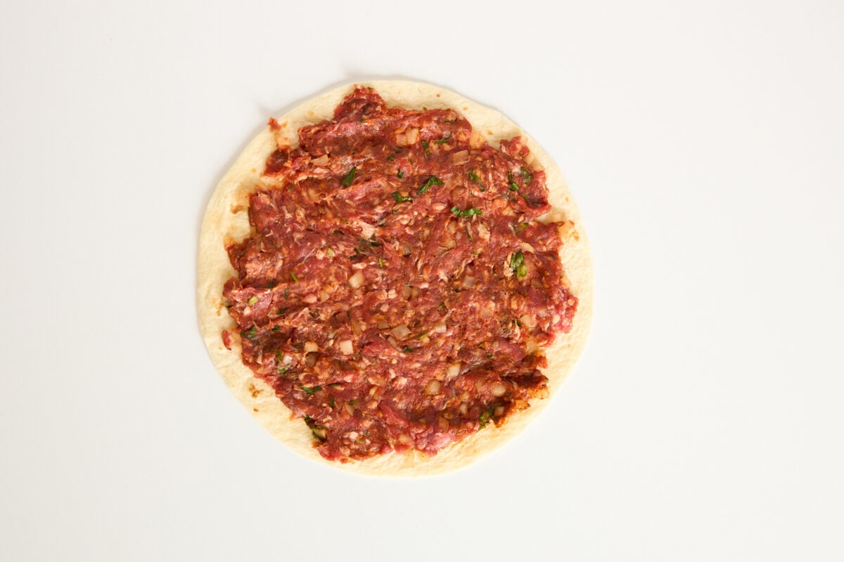 A round piece of flatbread topped with a raw, finely minced meat mixture, spread evenly to the edges, on a plain white background.