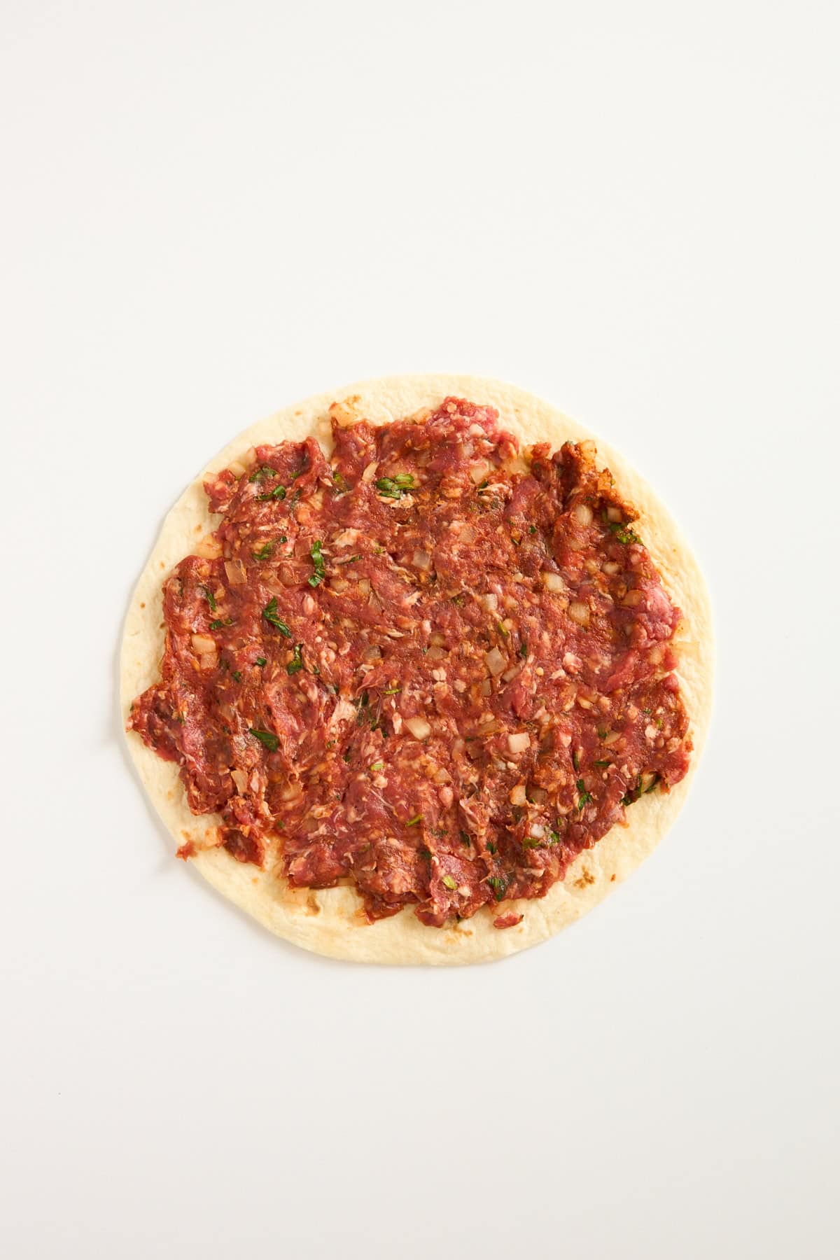 A round piece of flatbread topped with a raw, finely minced meat mixture, spread evenly to the edges, on a plain white background.