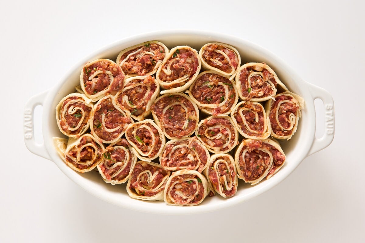A white oval baking dish filled with neatly arranged, uncooked meat pinwheels made from rolled dough with a visible minced meat and herb filling. The dish is placed on a plain white surface.