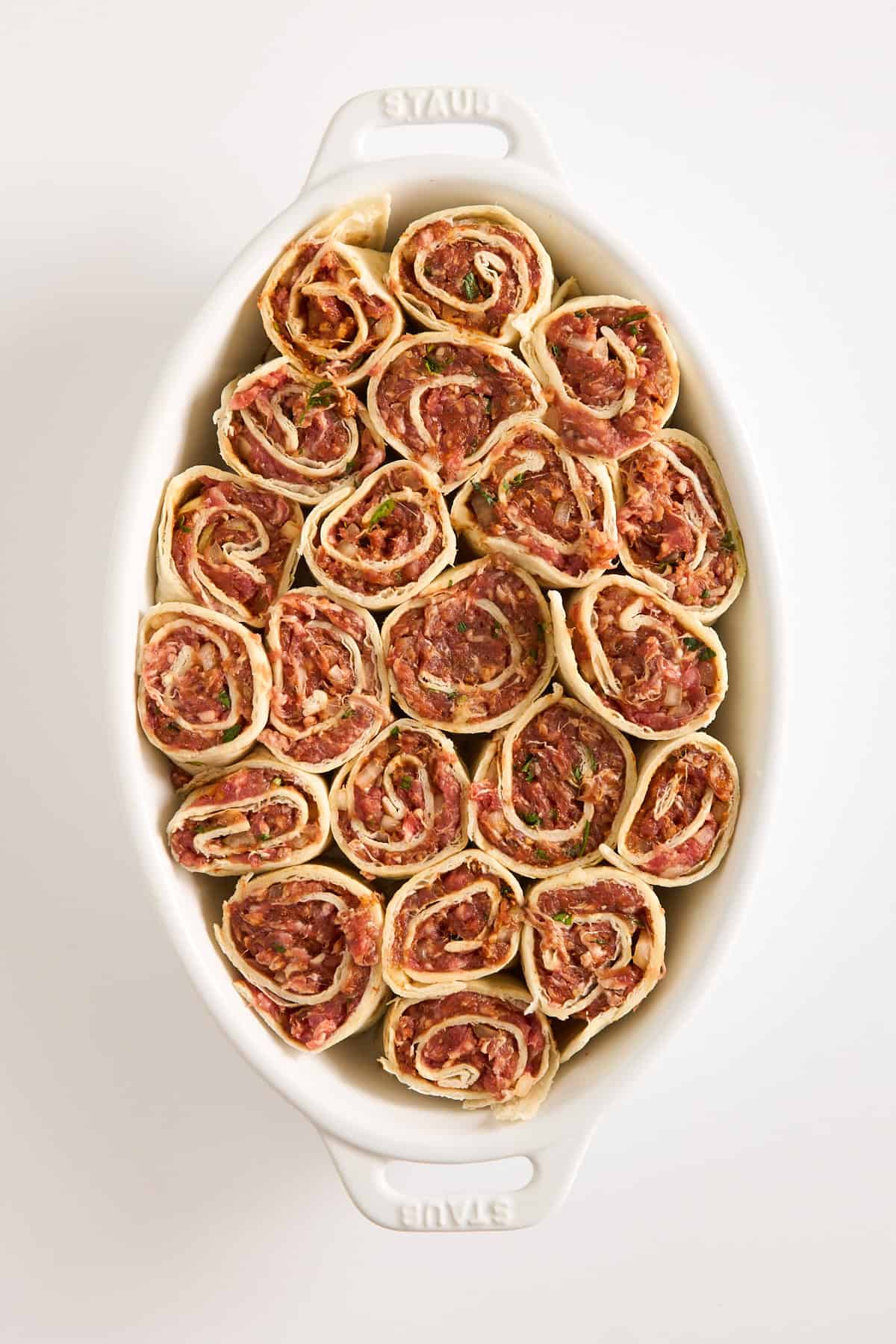 A white oval baking dish filled with neatly arranged, uncooked meat pinwheels made from rolled dough with a visible minced meat and herb filling. The dish is placed on a plain white surface.