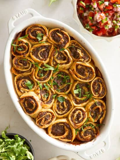 A white oval dish filled with golden-brown, spiral pinwheel rolls topped with chopped fresh herbs. Surrounding the dish are bowls of shredded lettuce, pico de gallo, sauce, and taco seasoning.