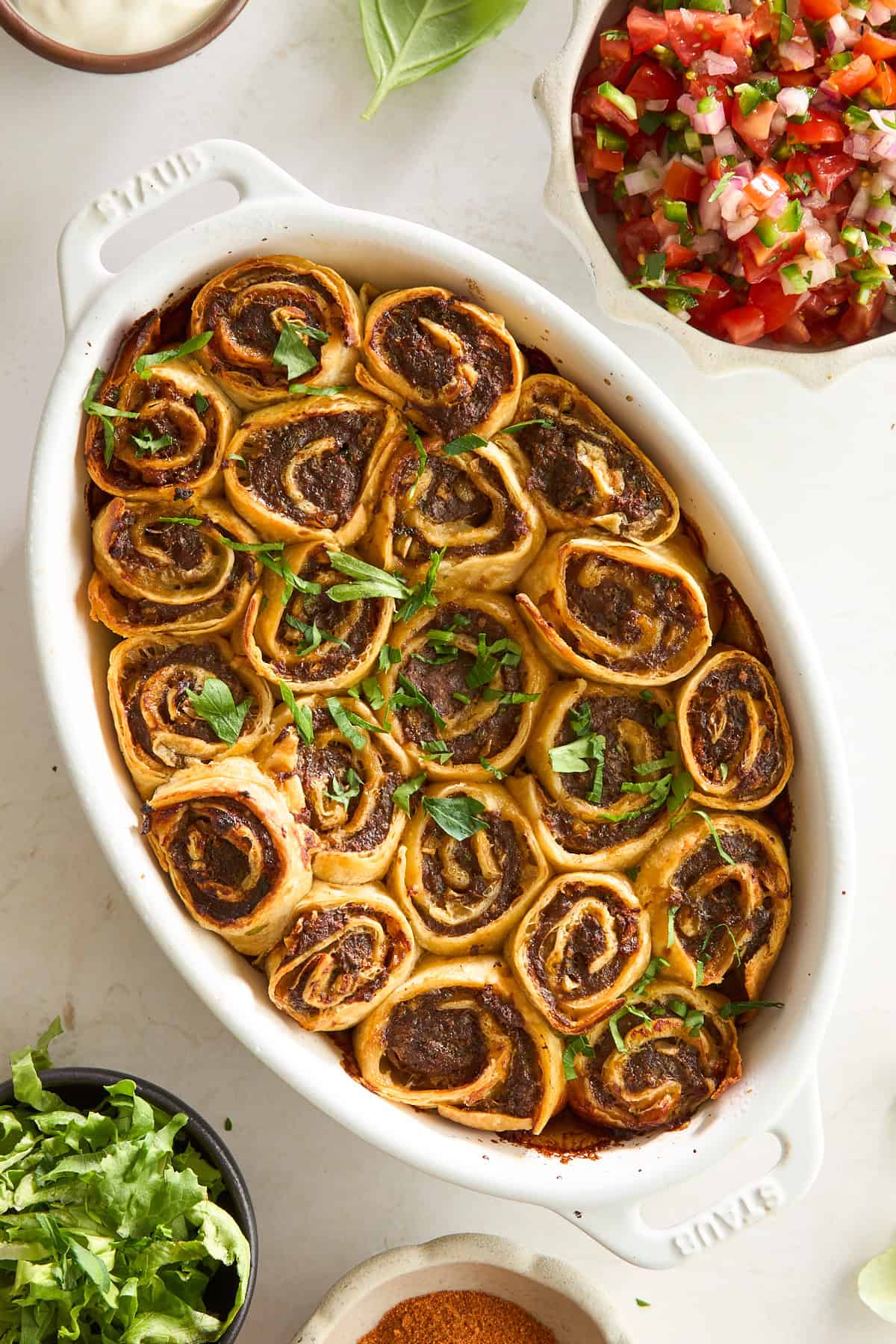 A white oval dish filled with golden-brown, spiral pinwheel rolls topped with chopped fresh herbs. Surrounding the dish are bowls of shredded lettuce, pico de gallo, sauce, and taco seasoning.