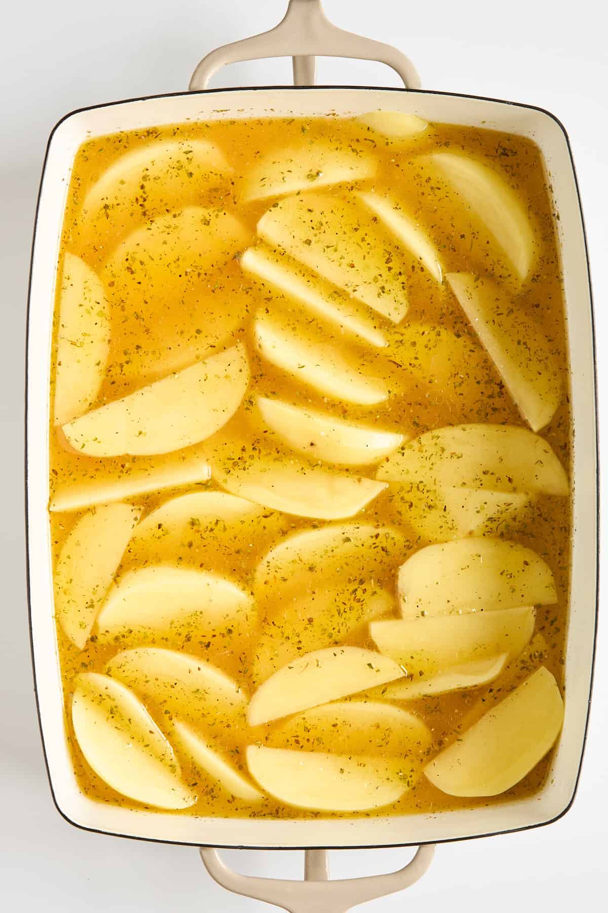 Sliced potatoes arranged in a white baking dish, covered in a yellow seasoned broth, ready to be cooked.