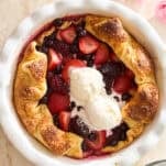 A rustic mixed berry galette with golden crust, filled with strawberries, blueberries, and blackberries, topped with two scoops of vanilla ice cream, on a light marble surface with pink flowers nearby.