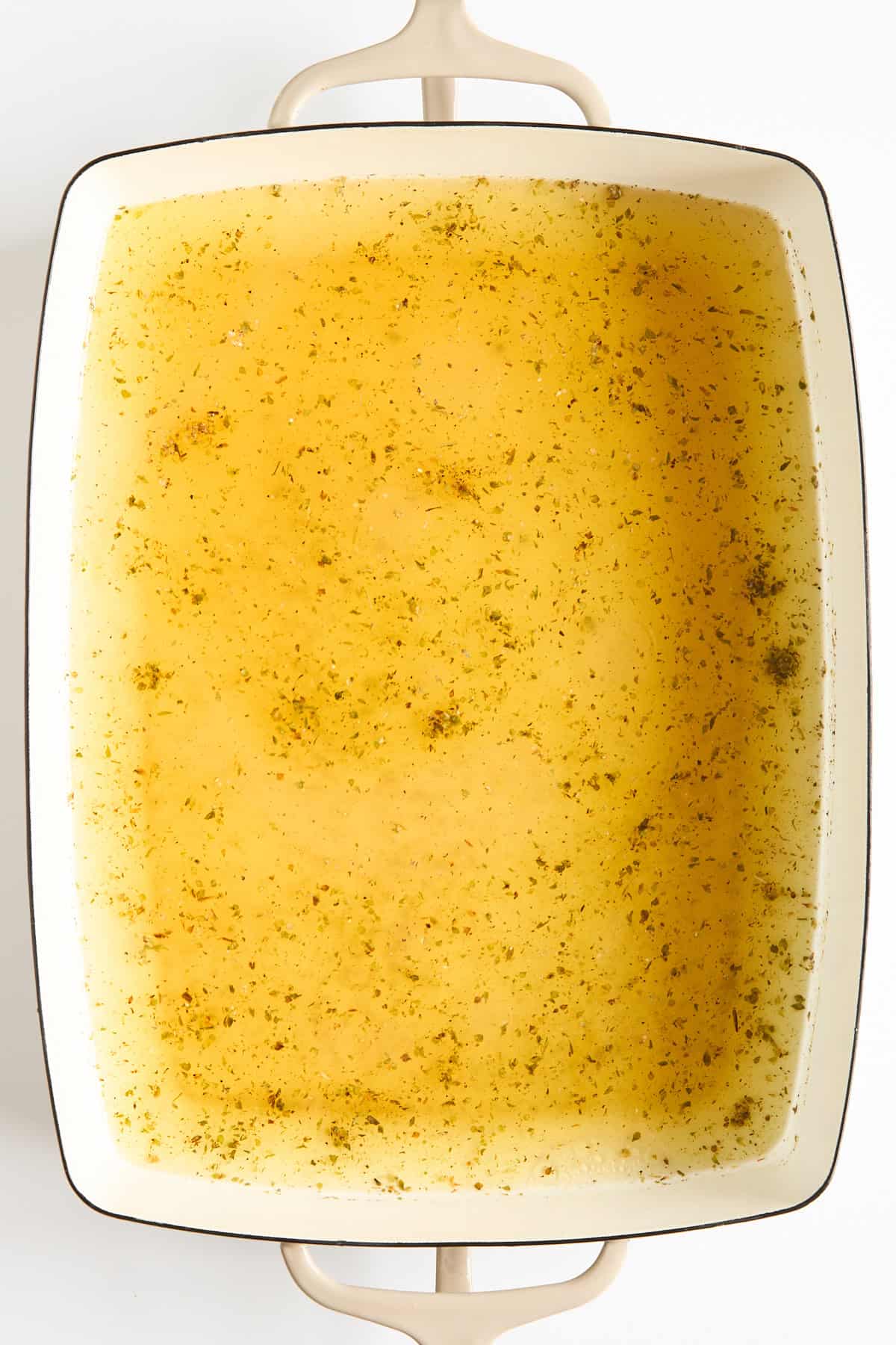 A rectangular white baking dish filled with a golden-yellow broth sprinkled with herbs, viewed from above. The dish has two handles and sits on a white surface.