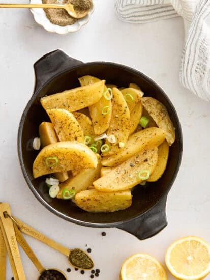A black dish filled with seasoned potato wedges garnished with sliced green onions, surrounded by measuring spoons with spices, lemon halves, and a striped napkin on a white surface.