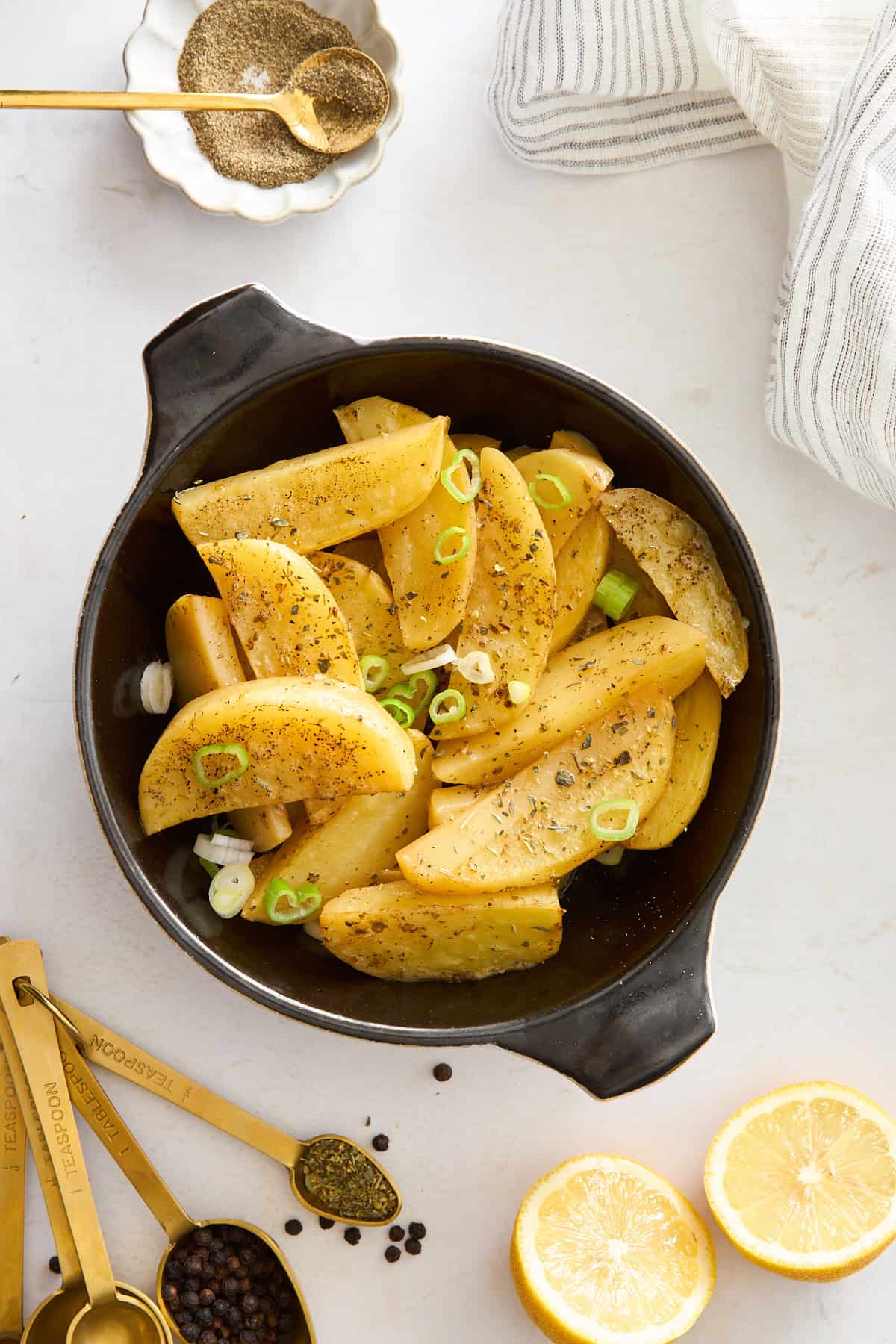 A black dish filled with seasoned potato wedges garnished with sliced green onions, surrounded by measuring spoons with spices, lemon halves, and a striped napkin on a white surface.