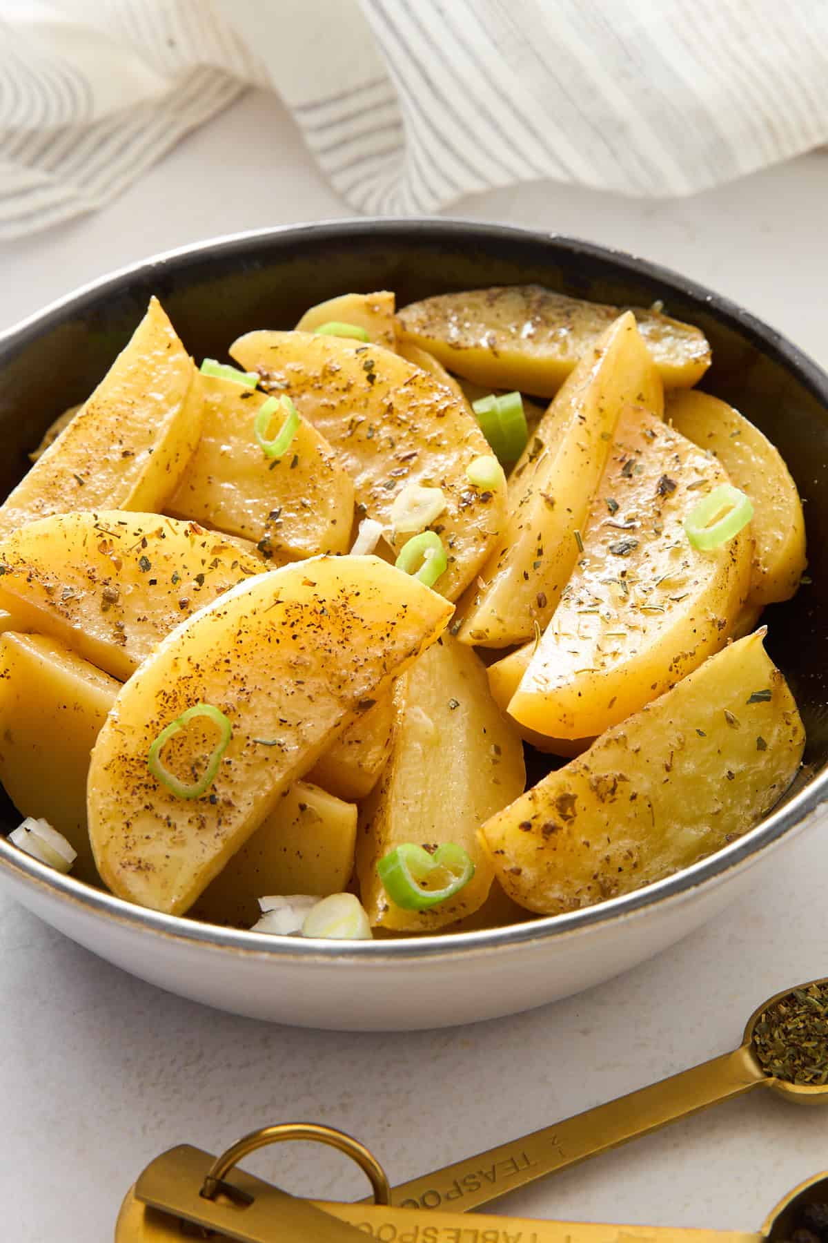 A bowl filled with seasoned potato wedges, garnished with chopped green onions and herbs, sits on a white surface next to golden measuring spoons and a striped cloth.