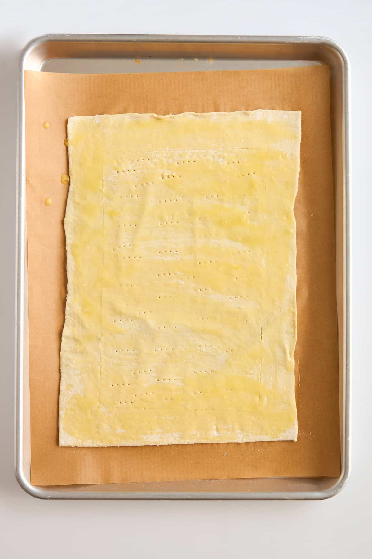 A rectangular sheet of dough brushed with egg wash and docked with fork marks sits on parchment paper on a baking tray.
