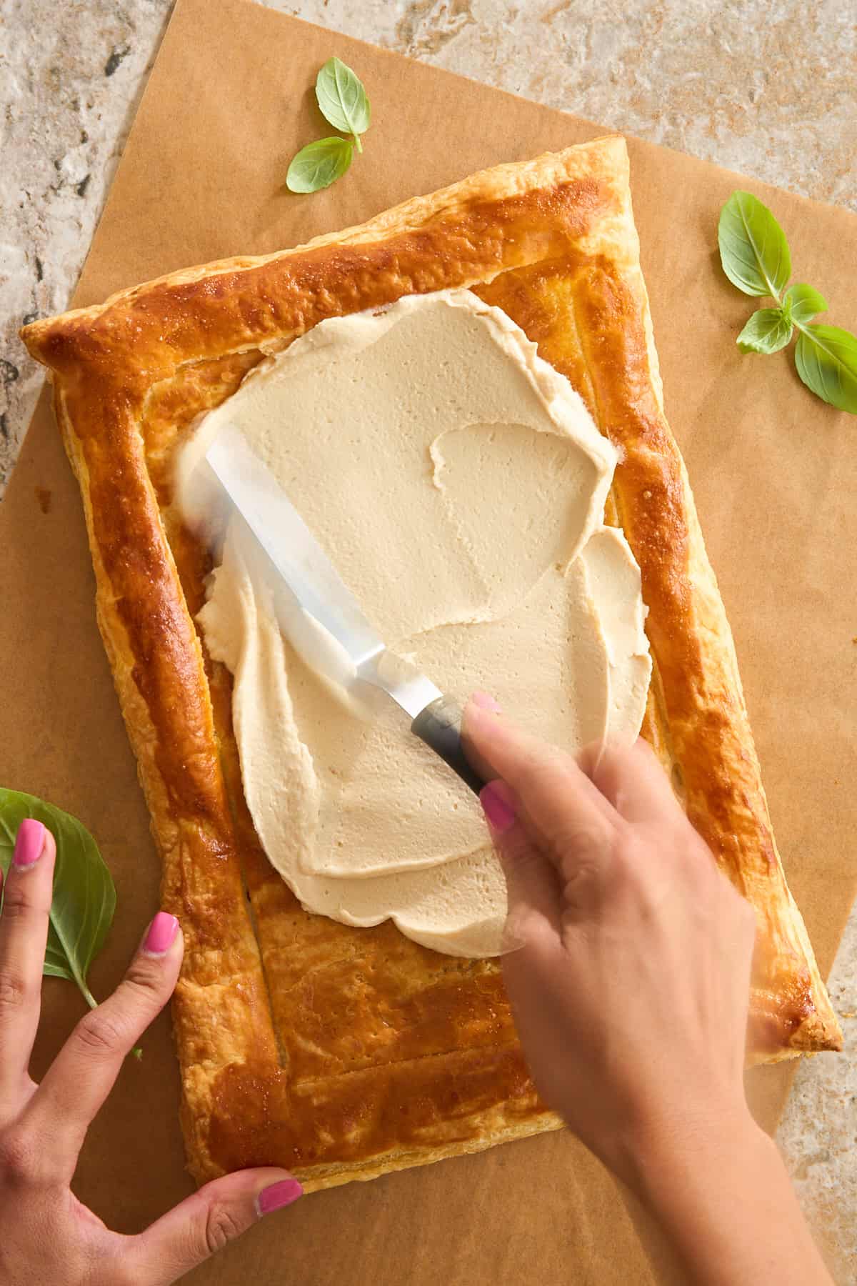 A person spreads a creamy, light-colored filling onto a rectangular, golden-brown puff pastry crust using an offset spatula. Fresh basil leaves and parchment paper are nearby.